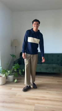Navy pullover with cream stripe, beige wide-leg trousers, and dark loafers. Indoor setting with a green couch and potted plants.