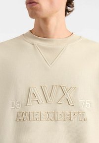 Felpa beige con colletto a coste, presenta il testo in rilievo "AVX 1975 AVIREX DEPT." con cuciture a contrasto, texture liscia, vestibilità morbida.