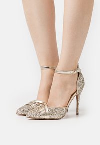 Glittering gold heels with a pointed toe, metallic accents, ankle straps, and textured glitter upper, featuring a stiletto heel.