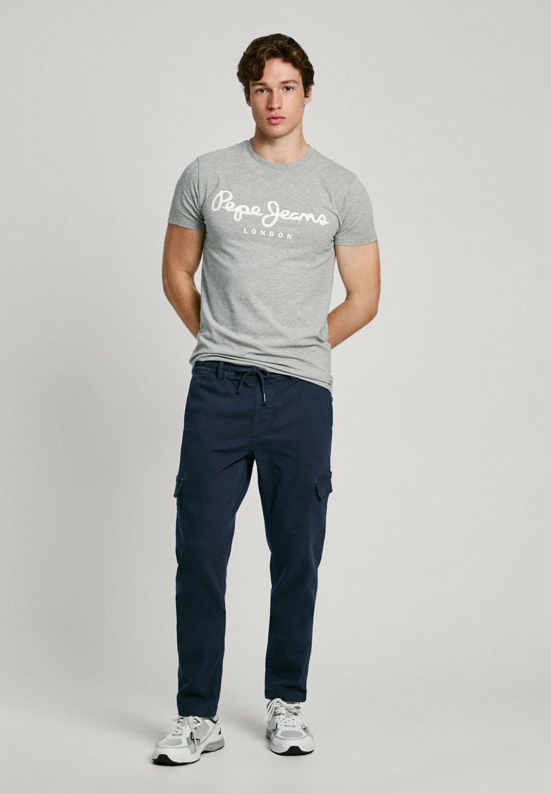 Noormees seisab otse, kannab halli Pepe Jeans T-särki, tumesiniseid cargopükse ja valgeid tosse ühevärvilise tausta ees.