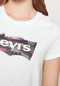 Biała bawełniana koszulka z dużym nadrukiem w kolorach fioletowym i zielonym, przedstawiającym logo „Levi's” w zakrzywionym obrysie.