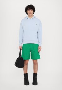 Sudadera azul claro con un bolsillo frontal y un logo negro, combinada con pantalones cortos verdes que tienen un gráfico blanco, una mochila negra y botas negras.