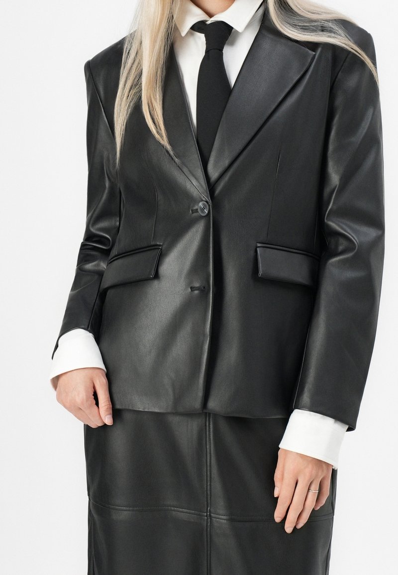 Femme portant un blazer en cuir noir et une jupe assortie avec des poignets de chemise blancs et une cravate noire, sur fond blanc uni.