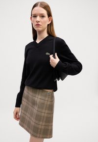 Maglione nero a scollo a V con logo, abbinato a una mini gonna a quadri in tonalità terra con accenti gialli. La modella tiene una borsa nera.