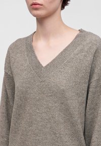 Personne portant un pull marron tricoté ample avec un col en V, montrant le cou et le haut de la poitrine sur un fond uni.