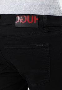 Gros plan sur la poche arrière et la ceinture d'un jean noir avec une étiquette de marque rouge et noire partiellement visible « HUGO » sur un fond de chemise grise.