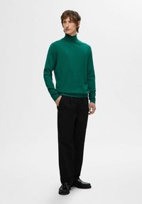 Zelen pulover s turtleneck ovratnikom, mehkega tekstila, oprijetega kroja. V kombinaciji z črnimi hlačami in črnimi loaferji, stoječ položaj pred nevtralno ozadjem.