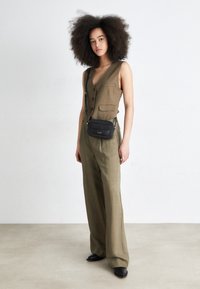 Oliwkowa lniana kamizelka z guzikami i kieszeniami, zestawiona z szerokimi oliwkowymi spodniami, czarną torbą na ramię i czarnymi butami.