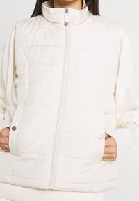 Biała kamizelka puchowa z wysokim kołnierzem, poziomym wzorem pikowania, zamkiem błyskawicznym z przodu oraz bocznymi kieszeniami z guzikami zatrzaskowymi. Teksturowany materiał.