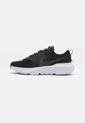 Black athletic shoe with mesh upper, grey accents, and a thick white speckled sole. Features a hook-and-loop strap and green heel tab.
