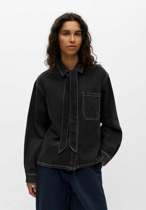 Femme aux longs cheveux bouclés portant une chemise en jean noire avec une poche avant et un pantalon sombre sur un fond blanc uni.