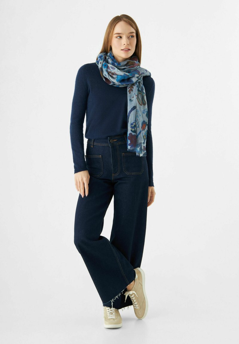 Haut à manches longues marine, jean large sombre et baskets beiges. L'écharpe bleue arbore un motif floral. Des teintes terreuses et une coupe détendue.