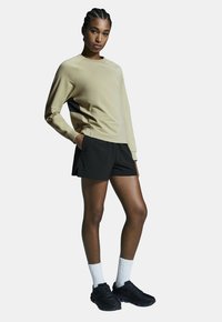 Beige lange mouwen shirt met zwarte zijkant, gecombineerd met zwarte shorts. Model draagt witte crew sokken en zwarte sportschoenen.