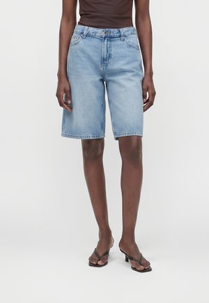 Persoon die lichtblauwe denim knielange shorts draagt, een donkerbruin mouwloos topje en zwarte sandalen met hakken, staand tegen een effen achtergrond.