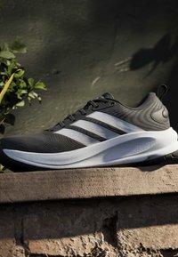 Baskets athlétiques noir et blanc avec trois bandes blanches sur le côté, posées sur une surface en pierre rugueuse sur un fond texturé sombre.