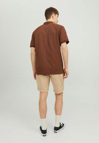 Polo shirt marron à manches courtes avec un motif texturé, doté d'un col et de manches raglan, porté avec un short beige retroussé et des baskets noires.