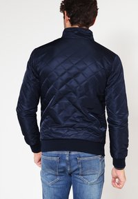 Mies, joka on pukeutunut tummansiniseen tilkkutakkiseen takkiin ja sinisiin farkkuihin, kääntyy pois yksinkertaista valkoista taustaa vasten.