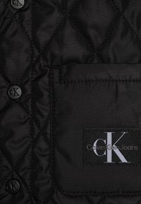 Giacca imbottita nera con bottoni rotondi neri, dotata di tasca frontale e targhetta con logo CK. Il materiale appare liscio e lucido.