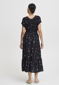Robe noire à motifs floraux avec des manches courtes, une taille smockée, un ourlet en couches et des motifs floraux multicolores. Associée à des sandales de couleur claire.