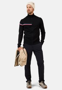 Jersey negro con cremallera y cuello alto, con una franja horizontal roja y azul. Combinado con pantalones de color marino y un gorro de punto beige.