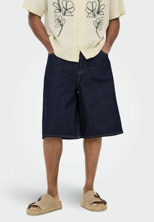 Hombre vistiendo pantalones cortos de mezclilla oscuros hasta la rodilla, sandalias beige con hebillas y una camisa clara con bordados florales negros, con las manos en los bolsillos.