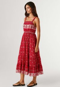Vrouw draagt een mouwloze rode midi-jurk met bloemenprint, brede bandjes en een gerimpelde rok, gecombineerd met bruine platte sandalen, staand tegen een effen achtergrond.
