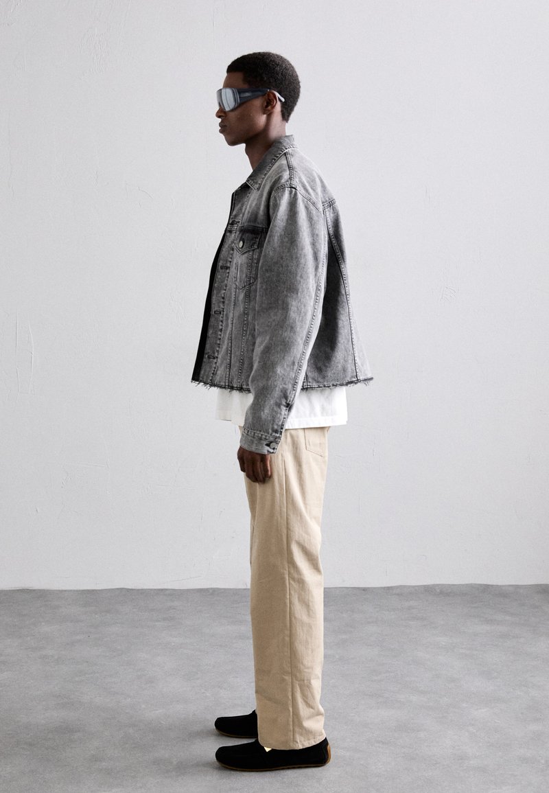 Gray denim jacket with frayed hem, black t-shirt, beige pants, and black shoes. Model wearing tinted sunglasses, standing against a plain backdrop.