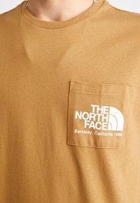 Brązowy bawełniany t-shirt z okrągłym dekoltem. Posiada kieszonkę na piersi z białym logo i napisem, gładką fakturę oraz standardowy krój.