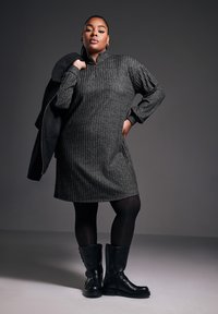 Vestito grigio scuro e textured con colletto a mezza zip, caratterizzato da un motivo a coste. Indossato con stivali neri до al ginocchio e una giacca nera appoggiata su un braccio.