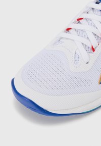 Chaussure de sport blanche avec empeigne en mesh, semelle en caoutchouc bleue et lacets d'accent rouge. Dispose d'une pointe lisse et arrondie et d'un design texturé.