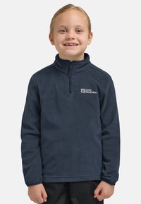 Bambino con capelli chiari che sorride, indossa un pullover in pile navy blue di Jack Wolfskin e pantaloni neri, in piedi su uno sfondo semplice.