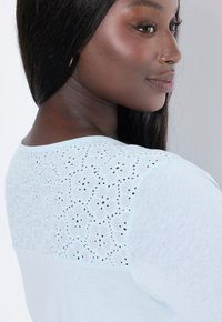 Světle modrý top s květinovou krajkou a detailem broderie anglaise v oblasti výstřihu a krátkými rukávy. Hladká textura a uvolněný střih.