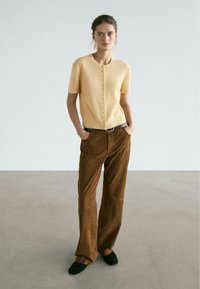 Short-sleeve, light yellow cardigan with a round neckline and button closure, paired with high-waisted brown corduroy pants. Black shoes.