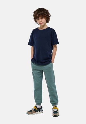 Garçon aux cheveux bouclés debout les mains dans les poches portant un t-shirt bleu marine, un jogging vert clair et des baskets multicolores sur fond blanc.