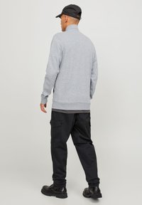 Siva zip-up dukserica s visokim ovratnikom, u kombinaciji s crnim cargo hlačama s bočnim džepovima i crnim čizmama. Glatka tekstura tkanine.