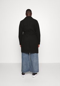 Zwarte lange jas met een ceintuur in de taille, voorzien van een brede kraag en een rechte snit. G gedragen over lichtblauwe wijde jeans en witte sneakers.