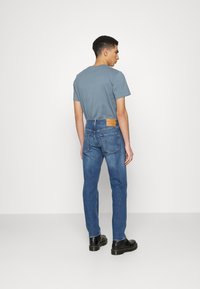 Jeans en denim bleu moyen avec une coupe régulière, dotés de deux poches arrière et d'un patch en cuir de marque. Assortis à un t-shirt gris à manches courtes.