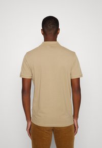 Polo em algodão bege com colarinho clássico e mangas curtas, apresentando um rótulo lateral, combinado com calças de veludo cotelê castanhas.