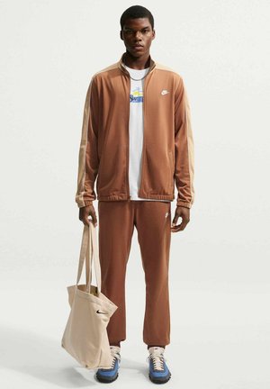 Mand iført brunt og beige Nike træningssæt, hvid t-shirt med grafik, blå og hvide sneakers, der holder beige mulepose med sort Nike-logo.
