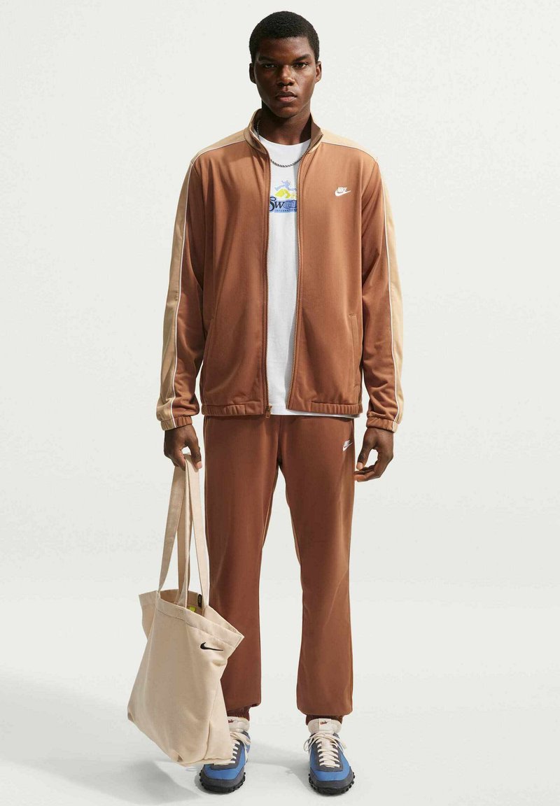 Mand iført brunt og beige Nike træningssæt, hvid t-shirt med grafik, blå og hvide sneakers, der holder beige mulepose med sort Nike-logo.