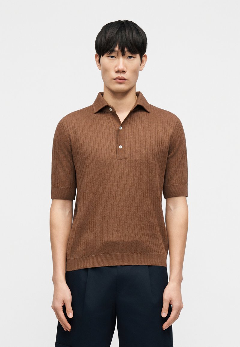 Polo marron en maille avec manches courtes, doté d'une patte de boutonnage à trois boutons et d'une texture côtelée, associé à un pantalon bleu marine foncé.