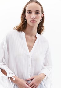 Weiße Bluse mit Knopfleiste, V-Ausschnitt, strukturiertem Stoff und elastischen Ärmeln mit Kordelzug-Akzenten, im lässigen Schnitt.