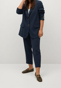 Mujer con blazer azul marino y pantalones recortados a juego, con mocasines verde oliva, de pie con una mano cerca de su cabeza.