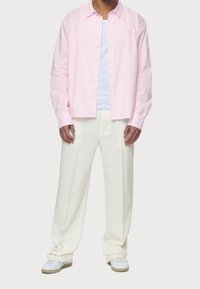 Różowa w paski koszula z długim rękawem, z zapięciem na guziki i kołnierzykiem, noszona na białym T-shircie. Zestawiona z beżowymi spodniami i białymi trampkami.