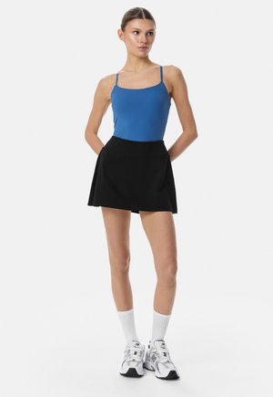 Žena stojící s rukama za zády, v modrém tílku, černé sukni, bílých kotníkových ponožkách a bílých sportovních teniskách.