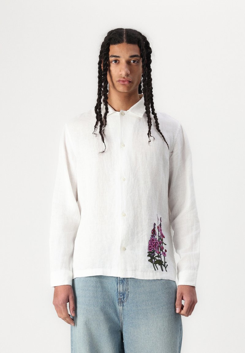 White linen button-up shirt with a collar, featuring purple floral embroidery on the left side, paired with light blue denim jeans.
