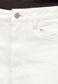 Λευκό τζιν denim σε κοντινό πλάνο που δείχνει κουμπί, θηλιά ζώνης, μπροστινή τσέπη και λεπτομέρειες με ανοιχτό μπεζ ραφές.