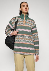 Fleece džemperis ar zaļu pamatu, ar ģeometriskiem rakstiem rozā, oranžā un baltā krāsā. Ar rāvējslēdzēju pie apkakles, garām piedurknēm un sānu kabatām.