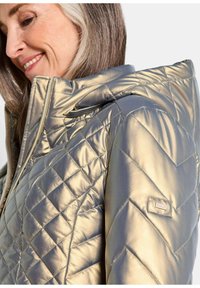 Goldene, gesteppten Jacke mit Kapuze, Reißverschluss und einer strukturierten, metallischen Oberfläche sowie einem Logo-Patch am Ärmel.