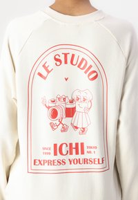Sudadera color crema con un gráfico vibrante en rojo que incluye una cámara y un personaje de dibujos animados, el texto dice "LE STUDIO" y "EXPRESS YOURSELF."